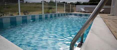 Una piscina con barandilla metálica de seguridad, rodeada por una valla y un área de descanso con sillas.