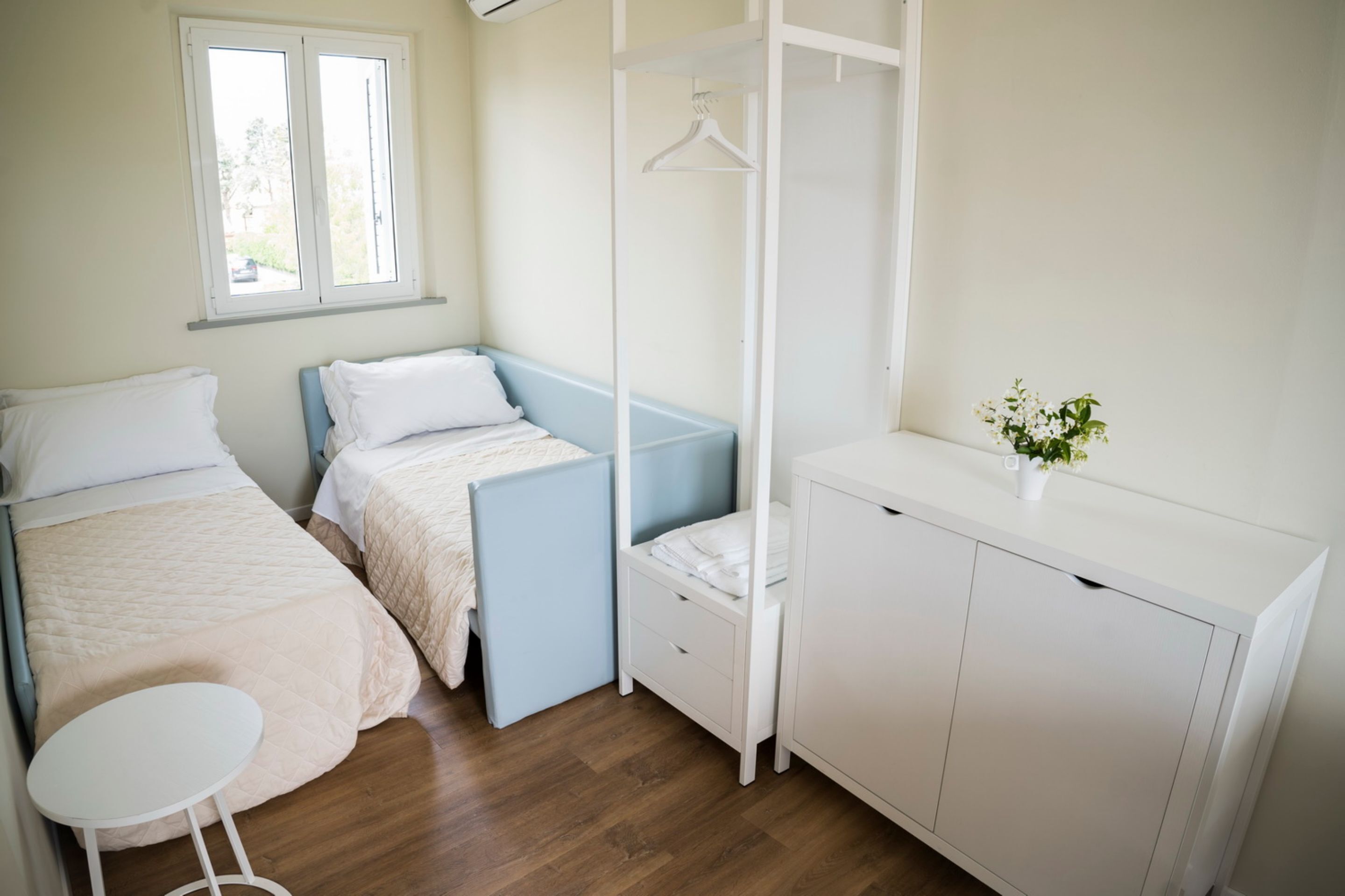 A bedroom with two beds, a white wardrobe, a small table, and a window.