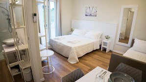 Una habitación de hotel moderna con una cama grande, un baño con ducha, una zona de comedor con una mesa preparada para dos personas y una mesita auxiliar con un florero.