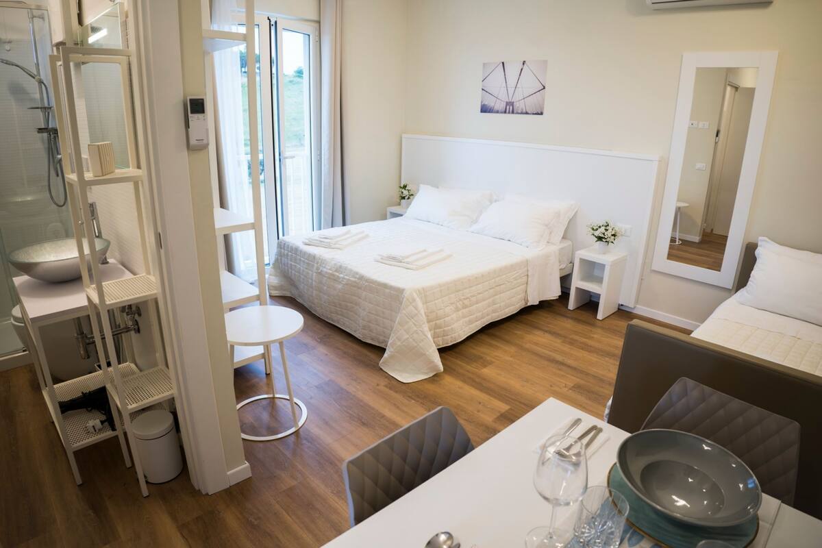 Une chambre d’hôtel moderne avec un grand lit, une salle de bain avec douche, un coin repas avec une table dressée pour deux personnes, et une petite table d’appoint avec un vase.