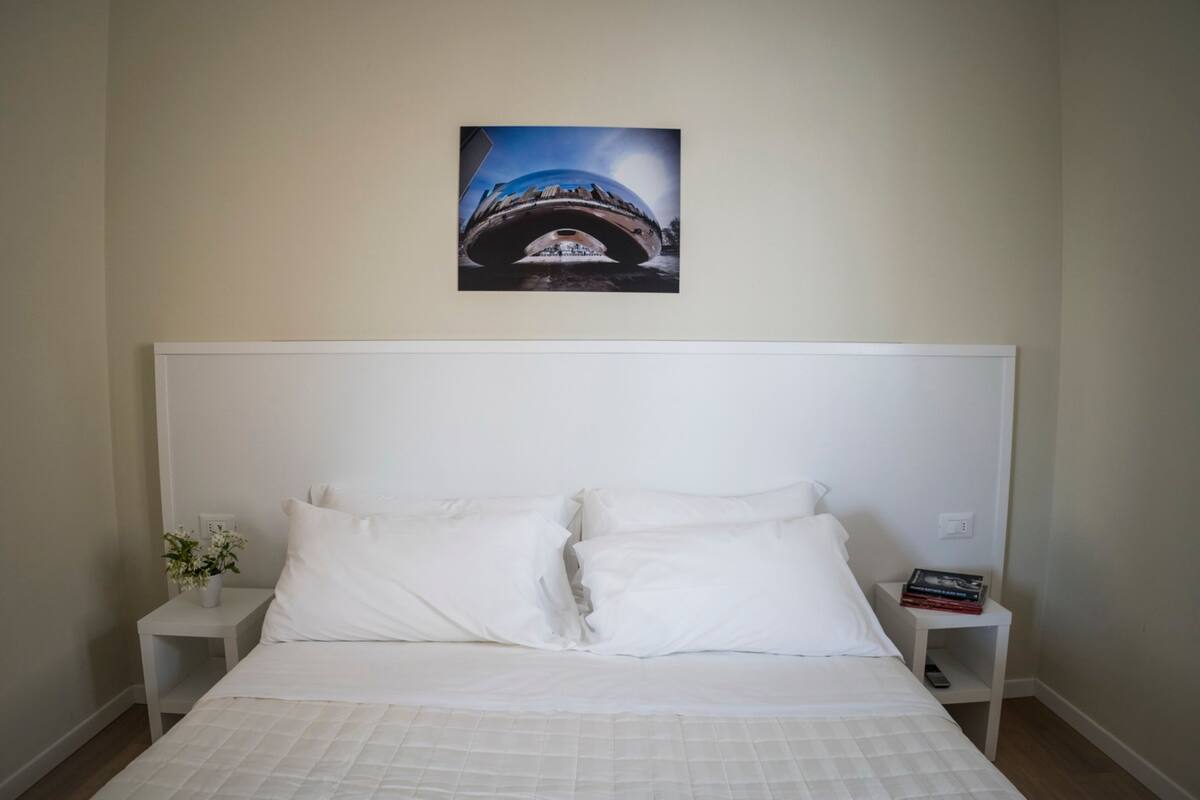 Une chambre à coucher avec un grand lit, une tête de lit blanche, deux tables de chevet avec des livres et un vase de fleurs, et un tableau encadré au mur.