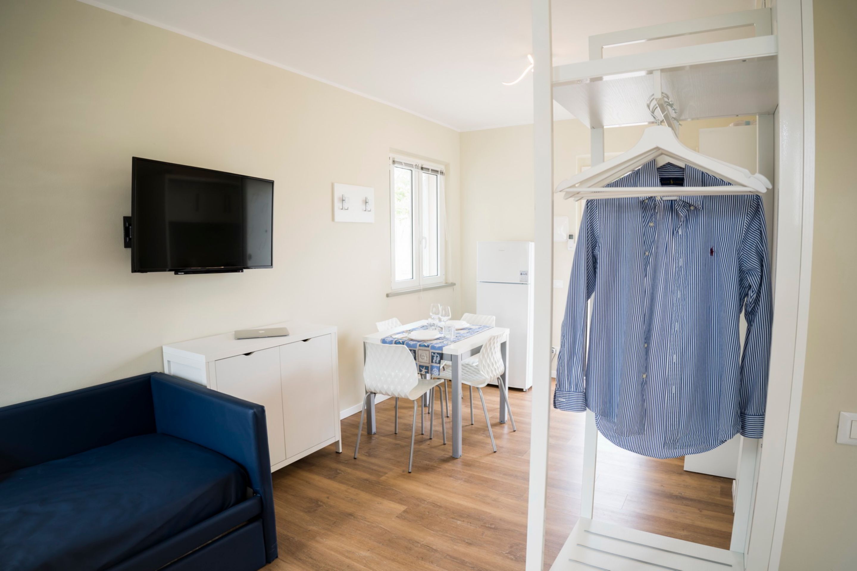 Un espace de vie compact comprenant un canapé, une table à manger avec des chaises, un téléviseur fixé au mur et un portant à vêtements.