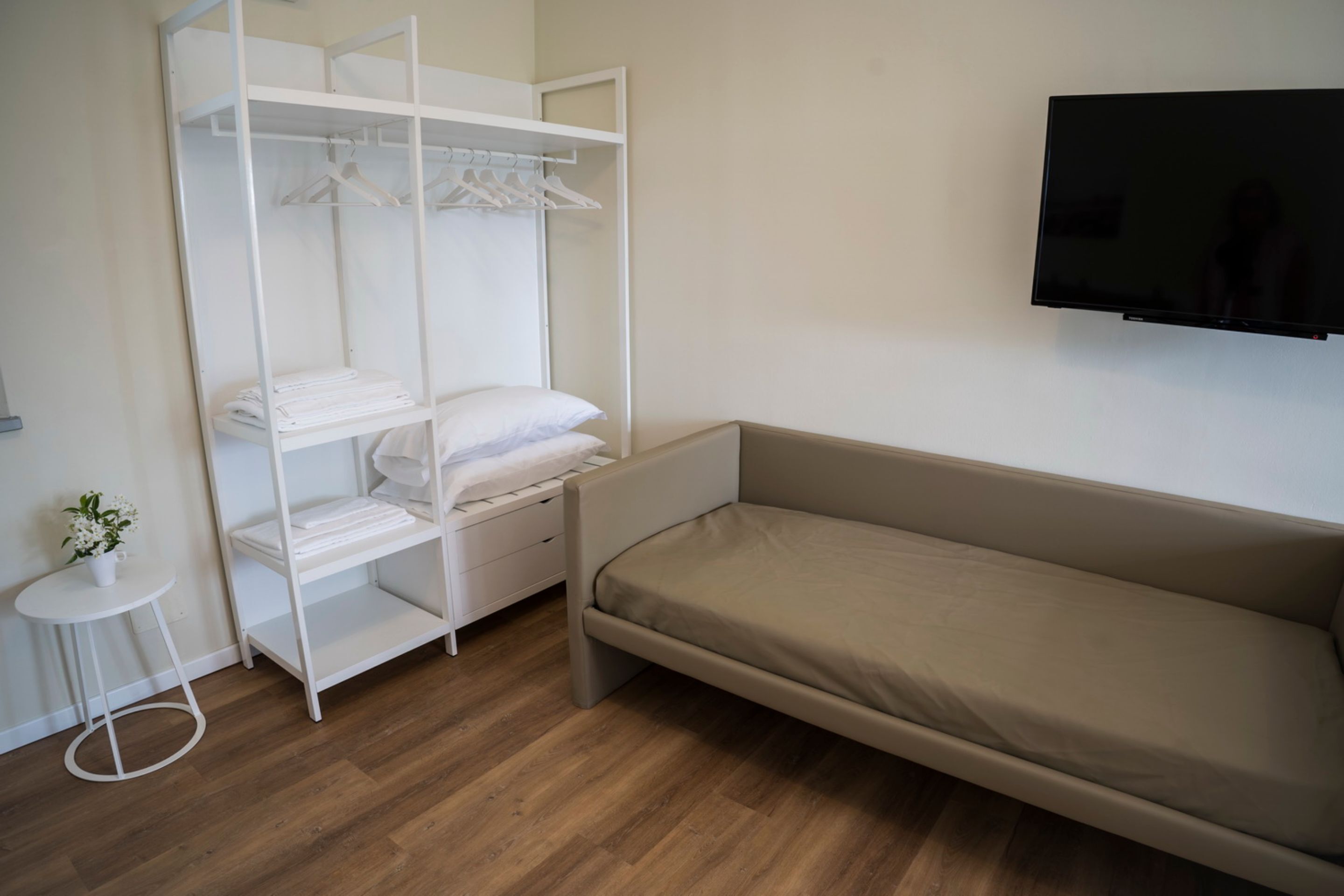 Ein Schlafzimmer mit Bett, Schrank, kleinem Tisch mit Pflanze und Fernseher an der Wand.