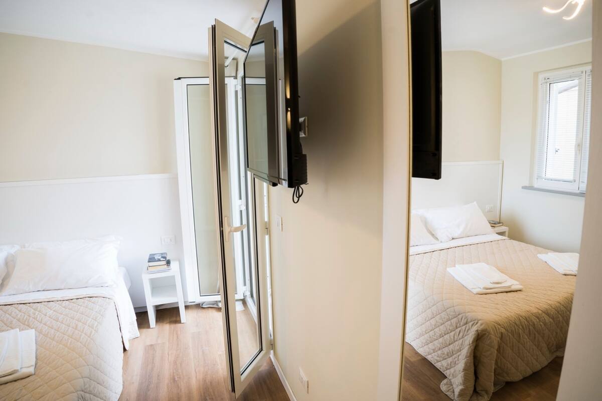 Une chambre d’hôtel avec deux lits, un téléviseur fixé au mur, une fenêtre avec des stores et une petite table de chevet sur laquelle se trouvent un livre et une lampe.
