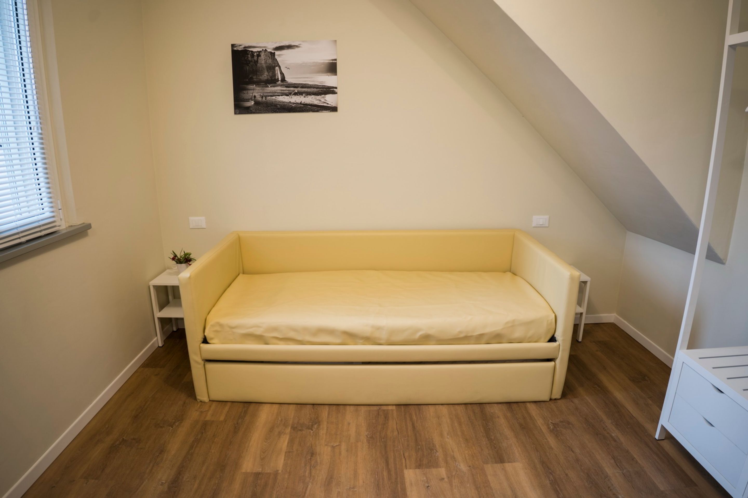 A small, single-bed room with a wooden floor, a yellow mattress, a small bedside table with a plant, and a picture on the wall.