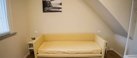 A small, single-bed room with a wooden floor, a yellow mattress, a small bedside table with a plant, and a picture on the wall.