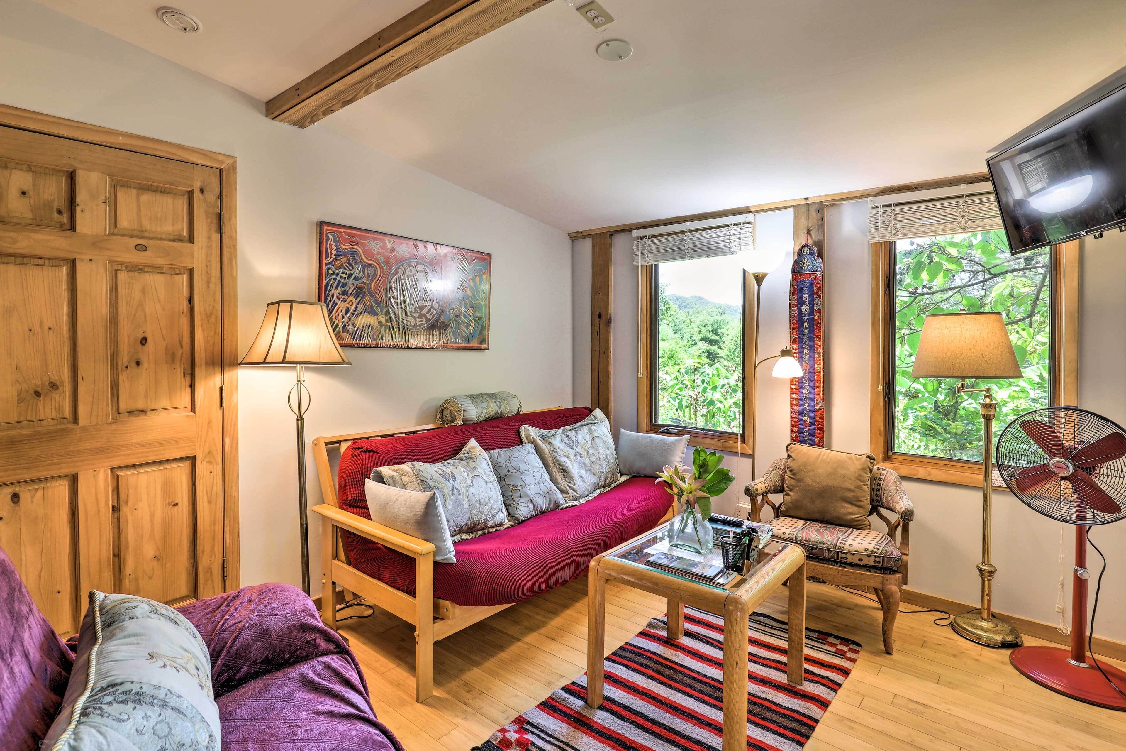 A cozy living room with a wooden sofa, a red armchair, and a coffee table. There is a painting on the wall, a window with a view of greenery, and a television mounted on the wall.