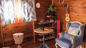 Ein Zimmer mit Holzwänden, einem runden Tisch mit Trommel, einem Stuhl mit floralem Kissen, einer Gitarre und einem Regal mit Pflanzen.