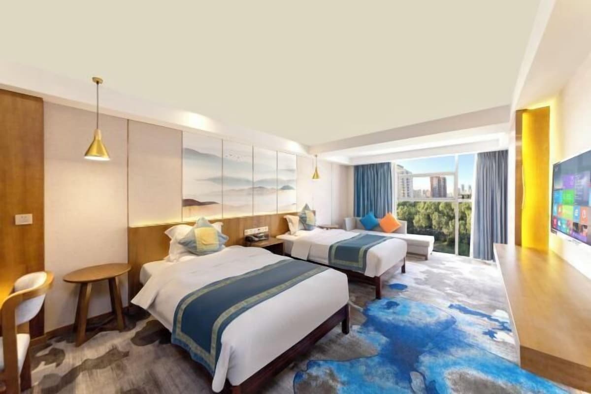 A modern hotel room with two beds, a desk, and a large window offering a city view.