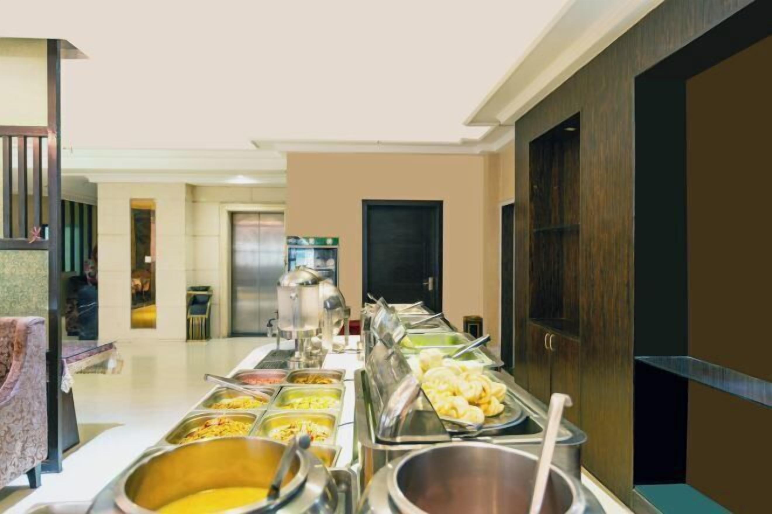 A buffet setup with various food items in metal containers, a coffee machine, and a seating area with a patterned tablecloth.