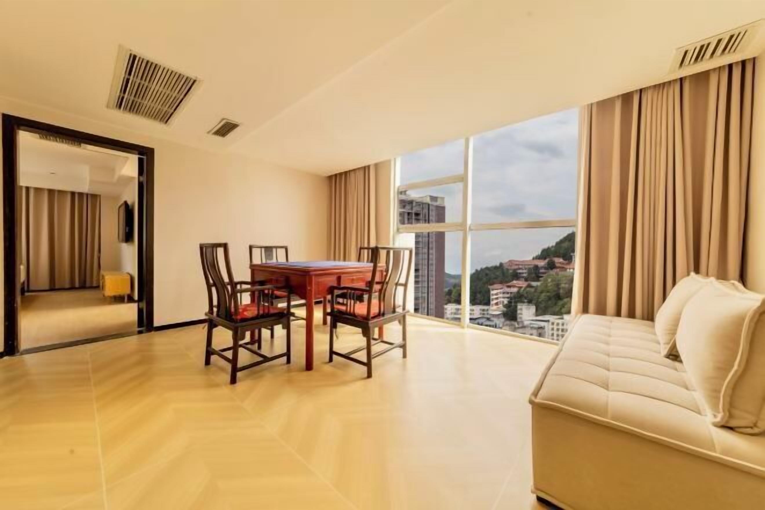 A modern hotel room with a dining table, chairs, a sofa, and a large window with a city view.