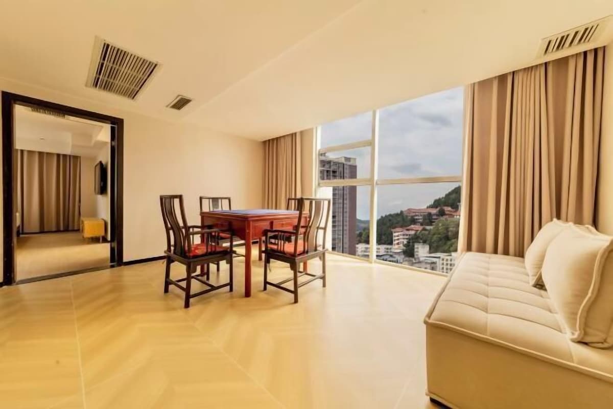 A modern hotel room with a dining table, chairs, a sofa, and a large window with a city view.