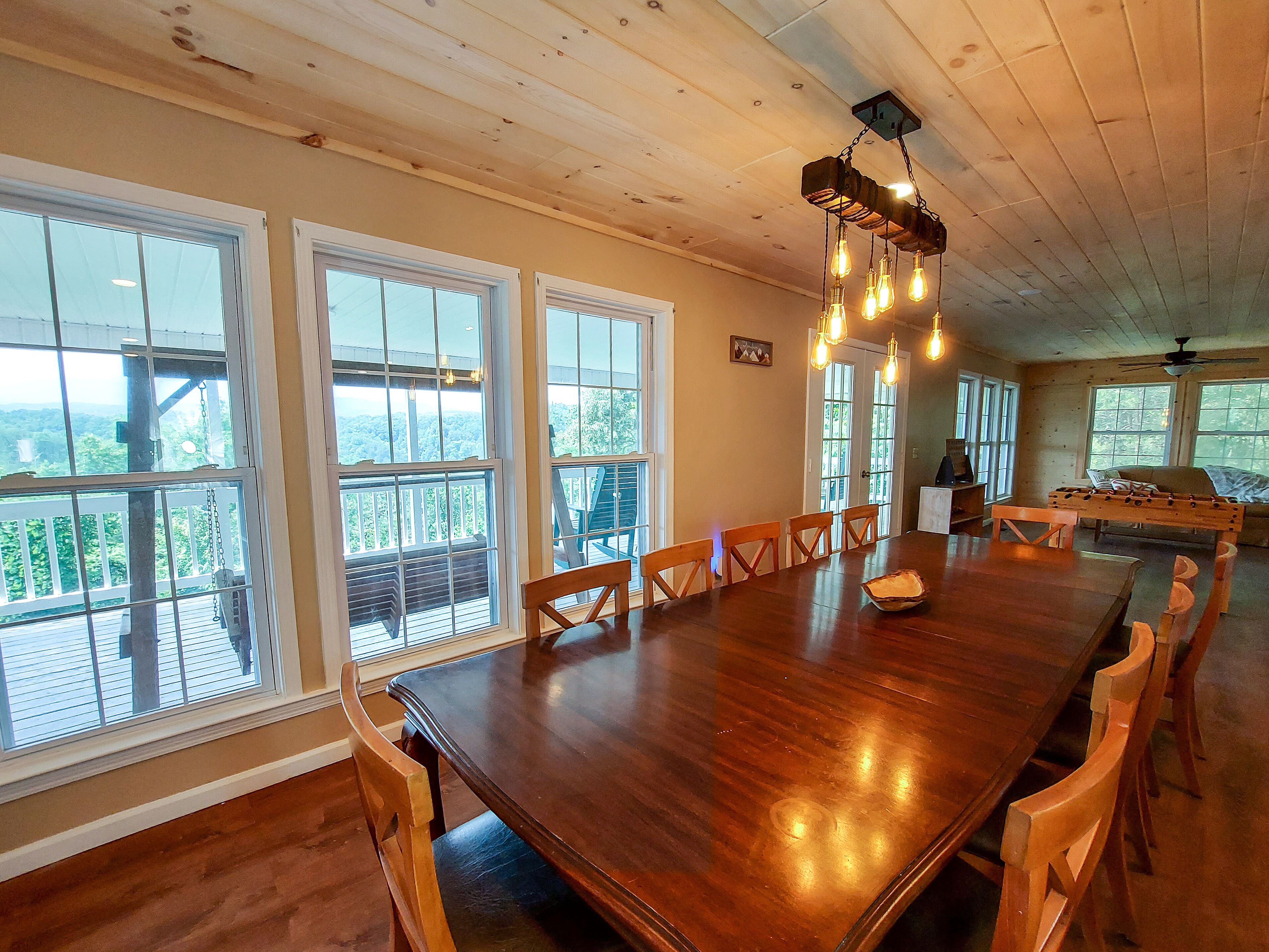 Un comedor amplio con una gran mesa de madera y sillas, ventanas grandes que ofrecen vistas del exterior y un ventilador de techo.