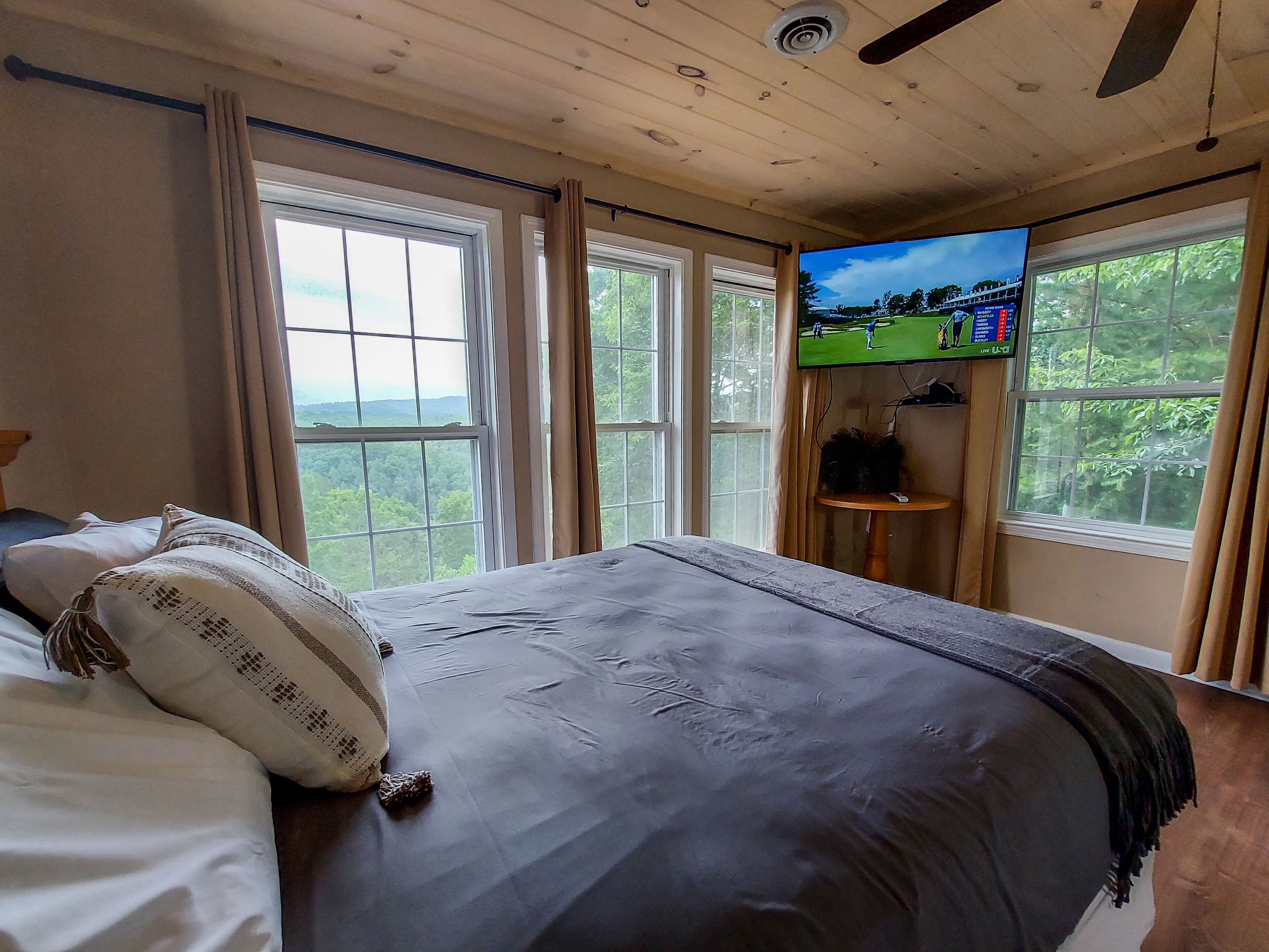 Ein Schlafzimmer mit einem Bett, großen Fenstern, einem Deckenventilator und einem Fernseher auf einem Tisch.