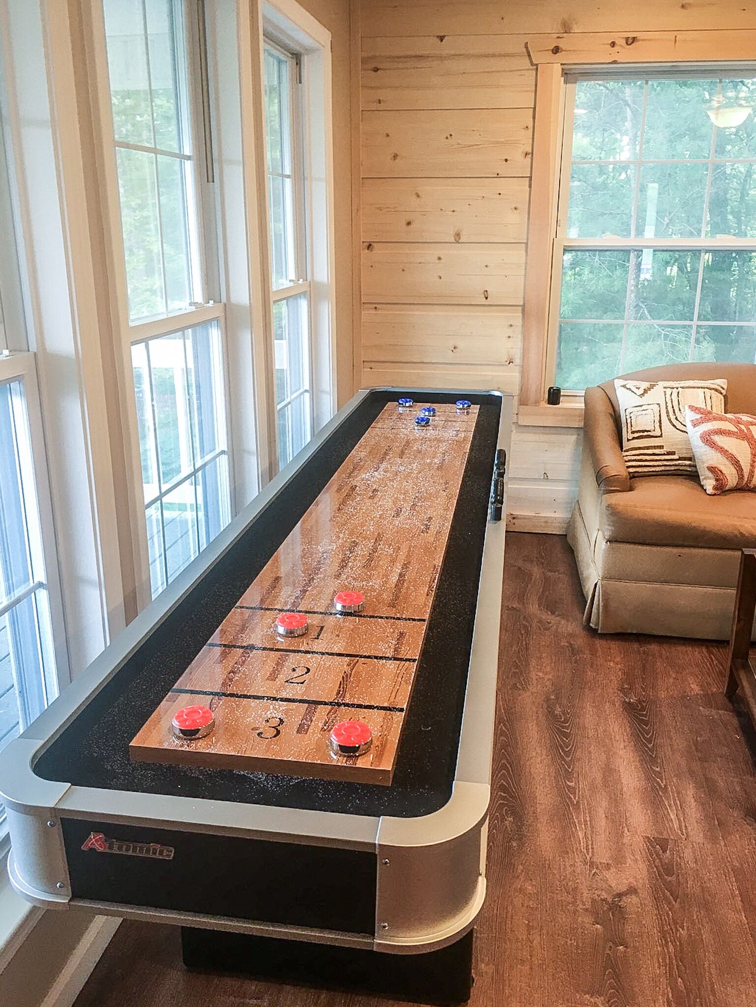 A shuffleboard table in a room with large windows and a couch.