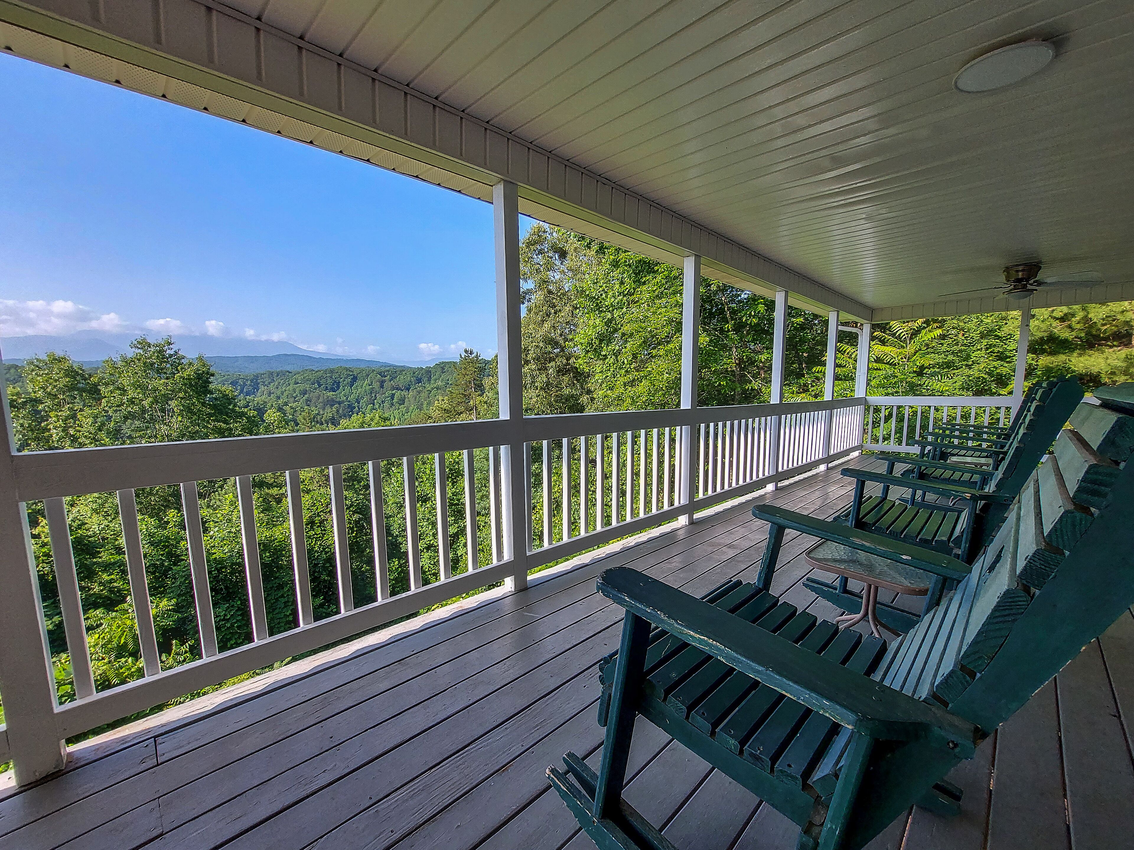 A covered porch with wooden railings and benches, overlooking a forested landscape with mountains in the distance.