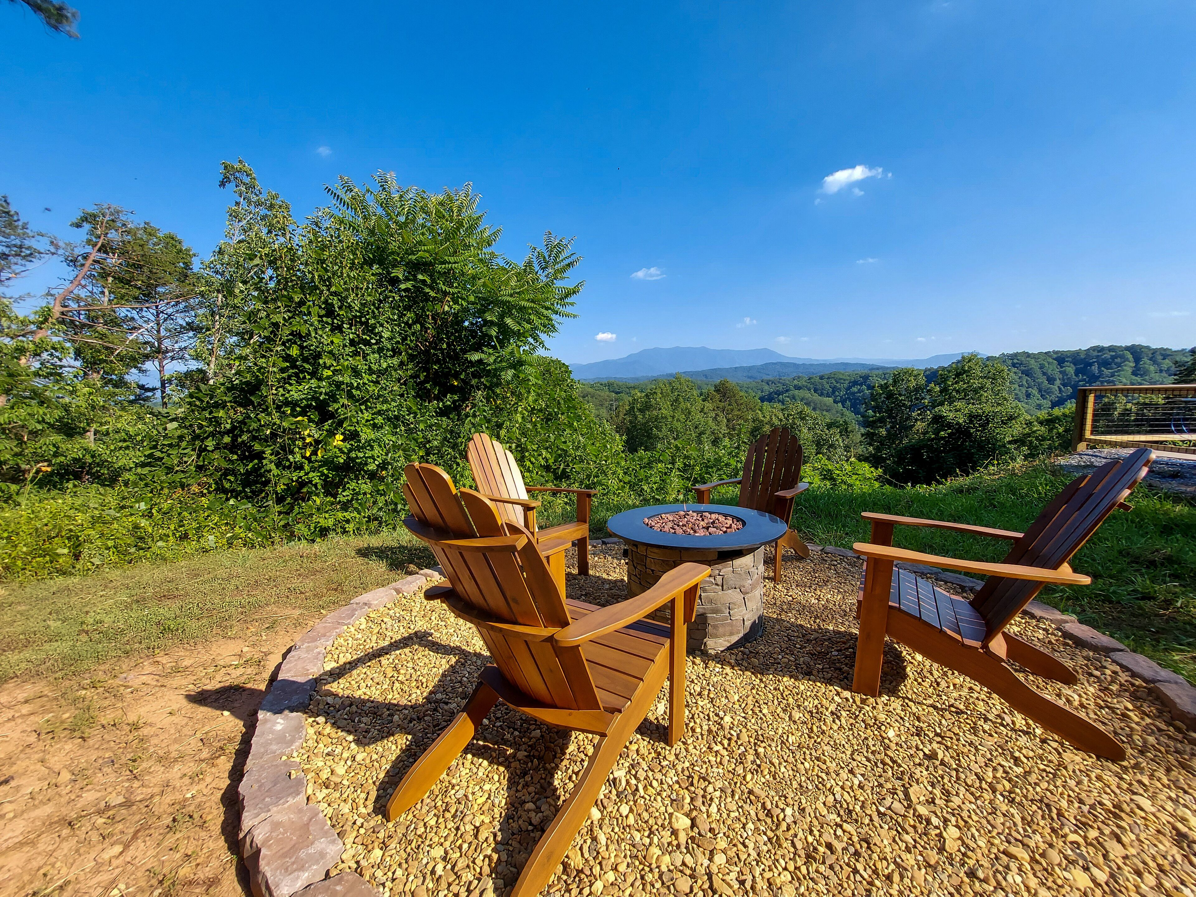 Una terraza de madera con sillas Adirondack y un pozo de fuego de piedra, rodeada por un camino de grava y abundante vegetación, con vistas a un paisaje montañoso.