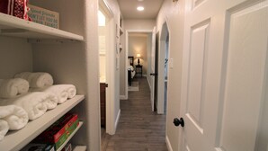 A narrow hallway with white walls, wooden flooring, and white doors. Shelves on the left hold neatly folded white towels and some books. A sign on the wall reads "Good friends don't let you do stupid things."