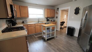 A kitchen with wooden cabinets, a stainless steel refrigerator, a gas stove, a coffee maker, and a small table with a cart.