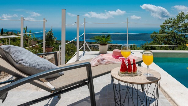 Un área de descanso junto a la piscina con vista al mar, una mesa con bebidas y sandía, y una silla de sol.