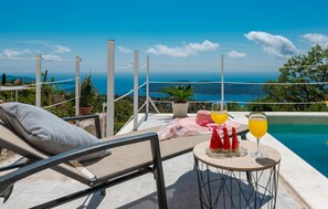 Un área de descanso junto a la piscina con vista al mar, una mesa con bebidas y sandía, y una silla de sol.