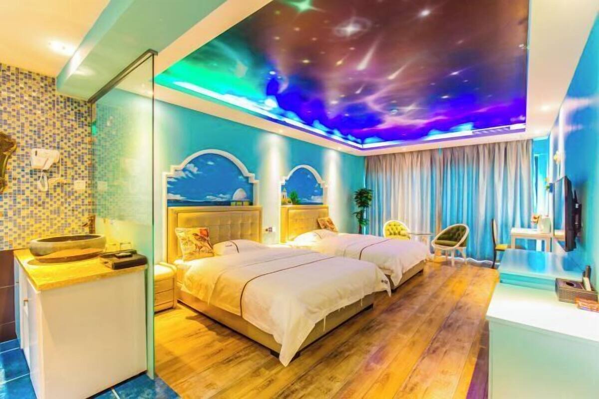 Habitación de hotel moderna con techo temático de galaxia, piso de madera, dos camas, tocador con espejo y vista a la ciudad.