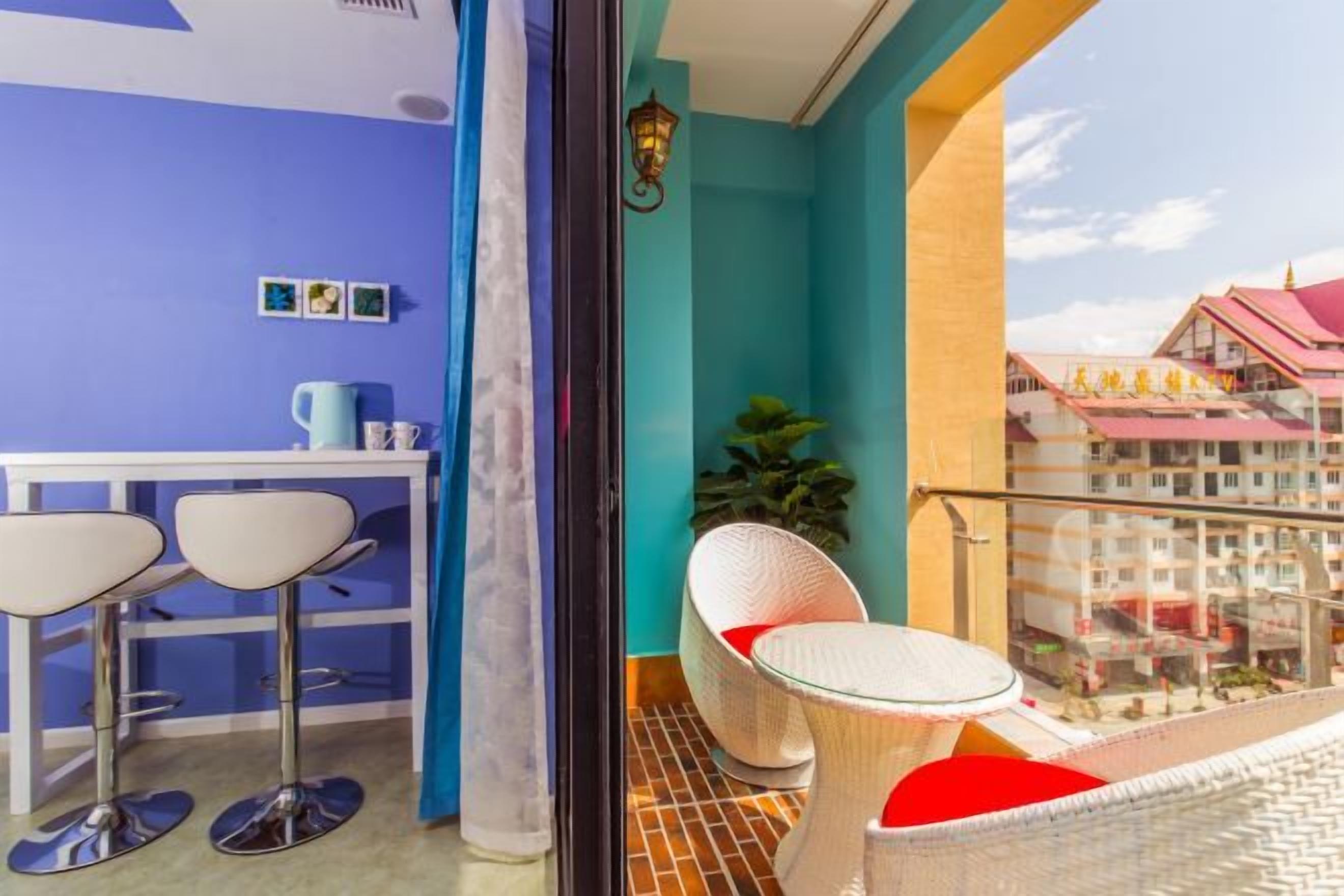 Ein Balkon mit Tisch und zwei Stühlen, eine kleine Küchenzeile mit Tisch und Hockern sowie Blick auf ein Gebäude mit roten Dächern.
