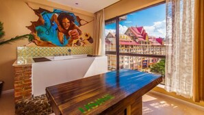 Una habitación con un ventanal grande, una mesa de madera con la inscripción 'LISKEEM', un mural en la pared y vistas de edificios coloridos al exterior.