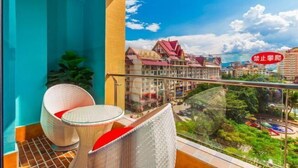 Un balcón con mobiliario de mimbre y una mesa de vidrio, con vista a un paisaje urbano con edificios y vegetación.