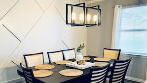 A dining area with a dark wooden table, six chairs, woven placemats, and a modern pendant light fixture. - Adversity Home - Open Concept, with Pacman Machine. Close to I-40 Airport (Amarillo)