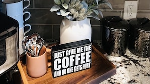A kitchen with a white cabinet containing a sugar container, a coffee maker, and a tray with a coffee mug and a sign that reads "Just give me the coffee and no one gets hurt." - Adversity Home - Open Concept, with Pacman Machine. Close to I-40 Airport (Amarillo)