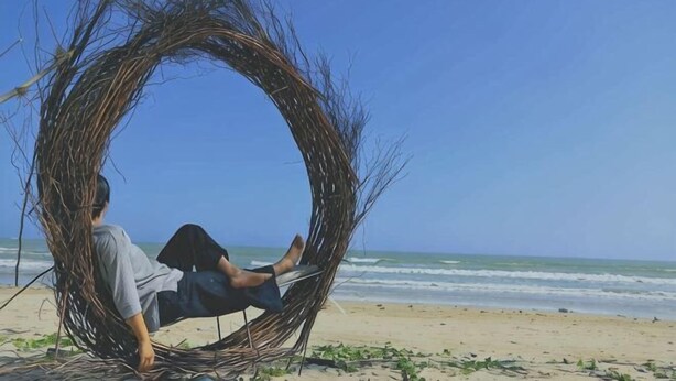 Une personne se détend dans une structure circulaire en osier sur une plage, avec l’océan en arrière-plan.