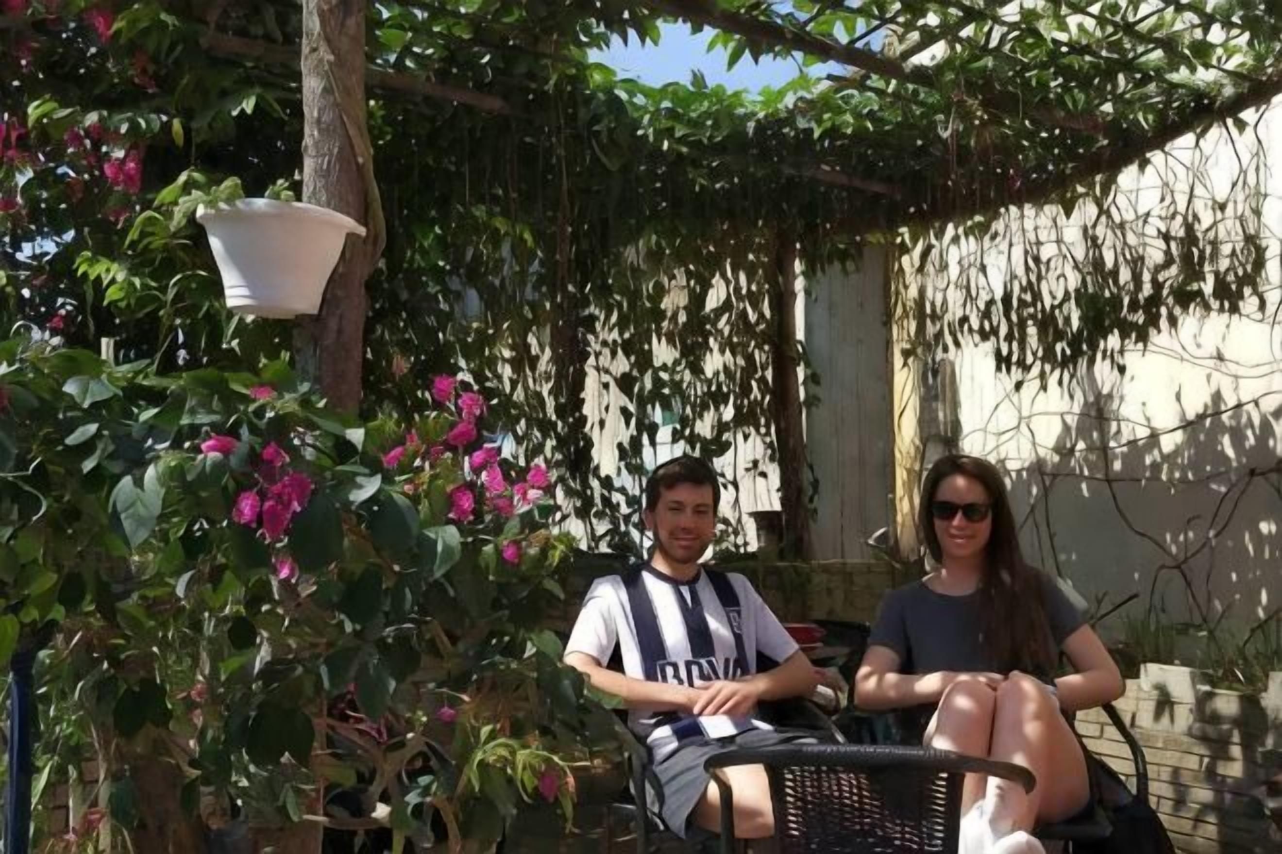 Two individuals seated on a bench under a pergola with hanging plants.