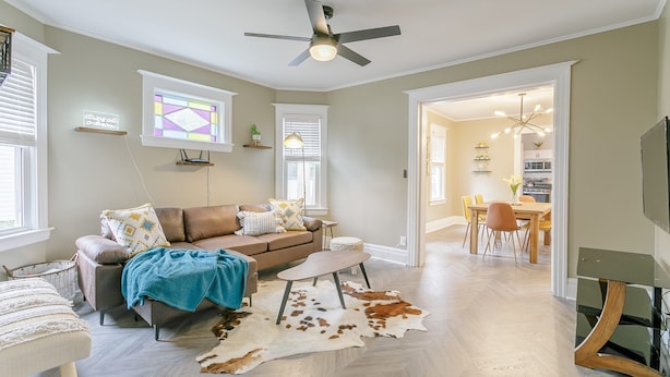 A modern living room with a brown leather sofa, a coffee table, and a cowhide rug. There is a ceiling fan, a TV stand, and a dining area visible in the background.