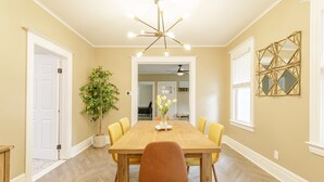 A dining room with a wooden table, six chairs, a chandelier, a potted plant, and a decorative mirror on the wall.
