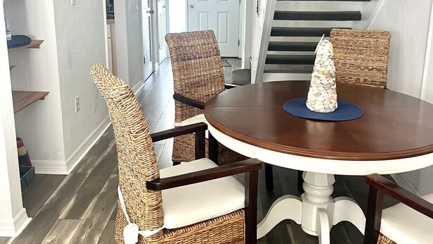 A dining area with wicker chairs and a round table, a staircase with black railings, and a white door in the background.