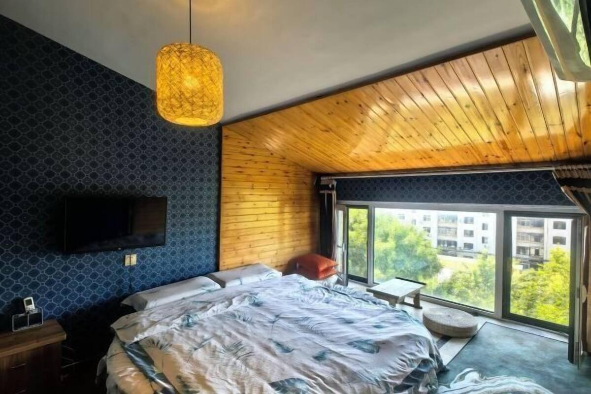 Un dormitorio moderno con una cama grande, un televisor montado en la pared y una ventana con vistas a edificios y árboles.