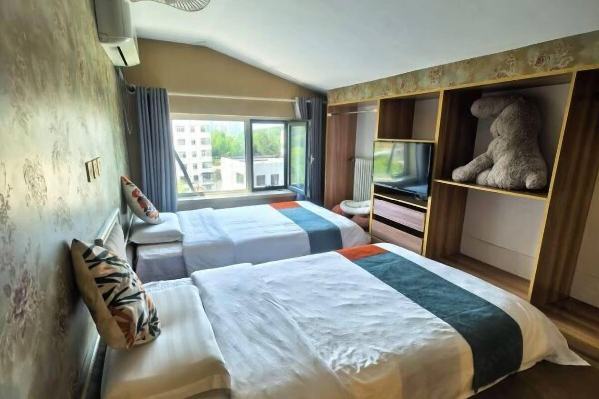 Habitación de hotel con dos camas, un ventanal grande, un osito de peluche en un estante y vistas a edificios.