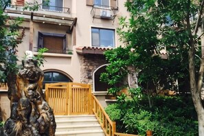 Un edificio con una escalera de madera que conduce a una entrada con reja, rodeado de vegetación y un árbol.