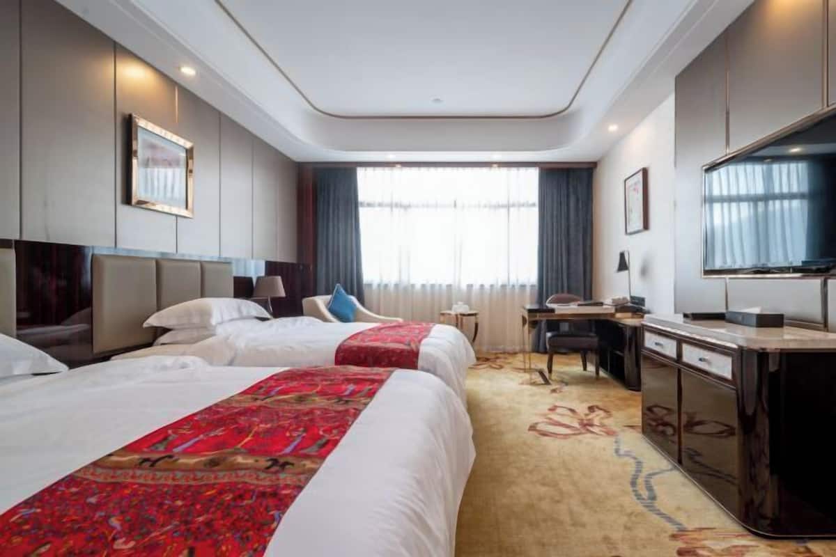 A hotel room with two beds, a desk, a television, and a large window with curtains.