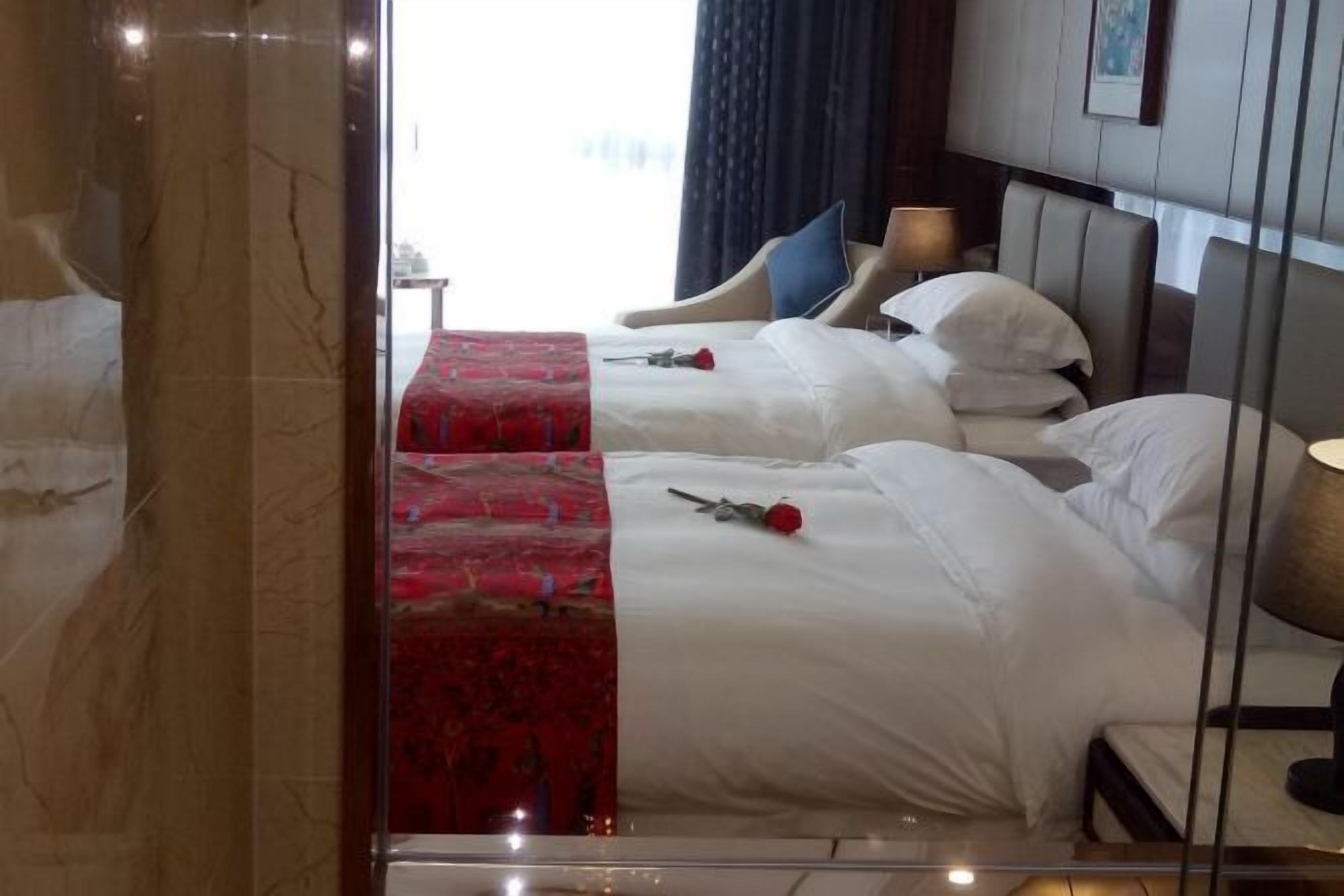 A hotel room with two beds, a red bed runner, white bedding, and a nightstand with a lamp.