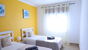 A hotel room with two beds, a window with sheer curtains, and artwork on the yellow wall. - 2 Bedroom Apartment in Portimão (Portimão)