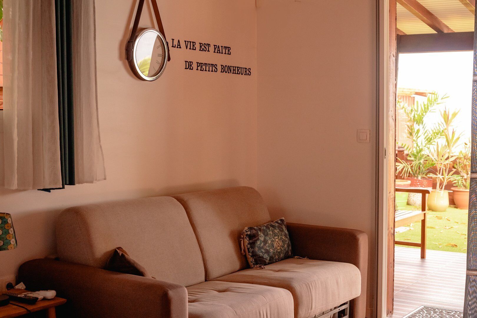 Habitación con sofá color beige, un reloj de pared y una puerta que da a un área exterior con plantas en macetas.