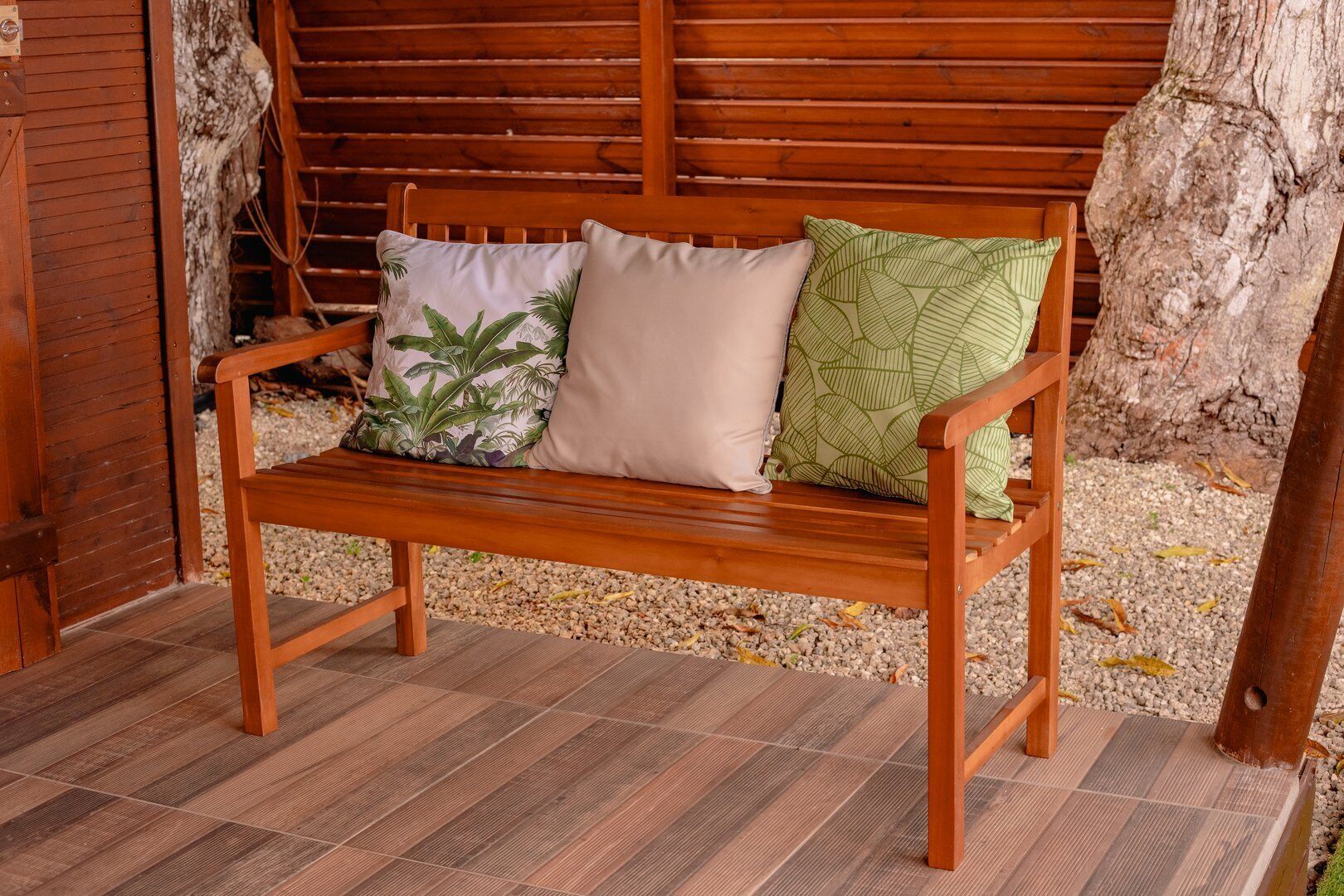 A wooden bench with three cushions against a wooden wall, surrounded by pebbles and a tree trunk.