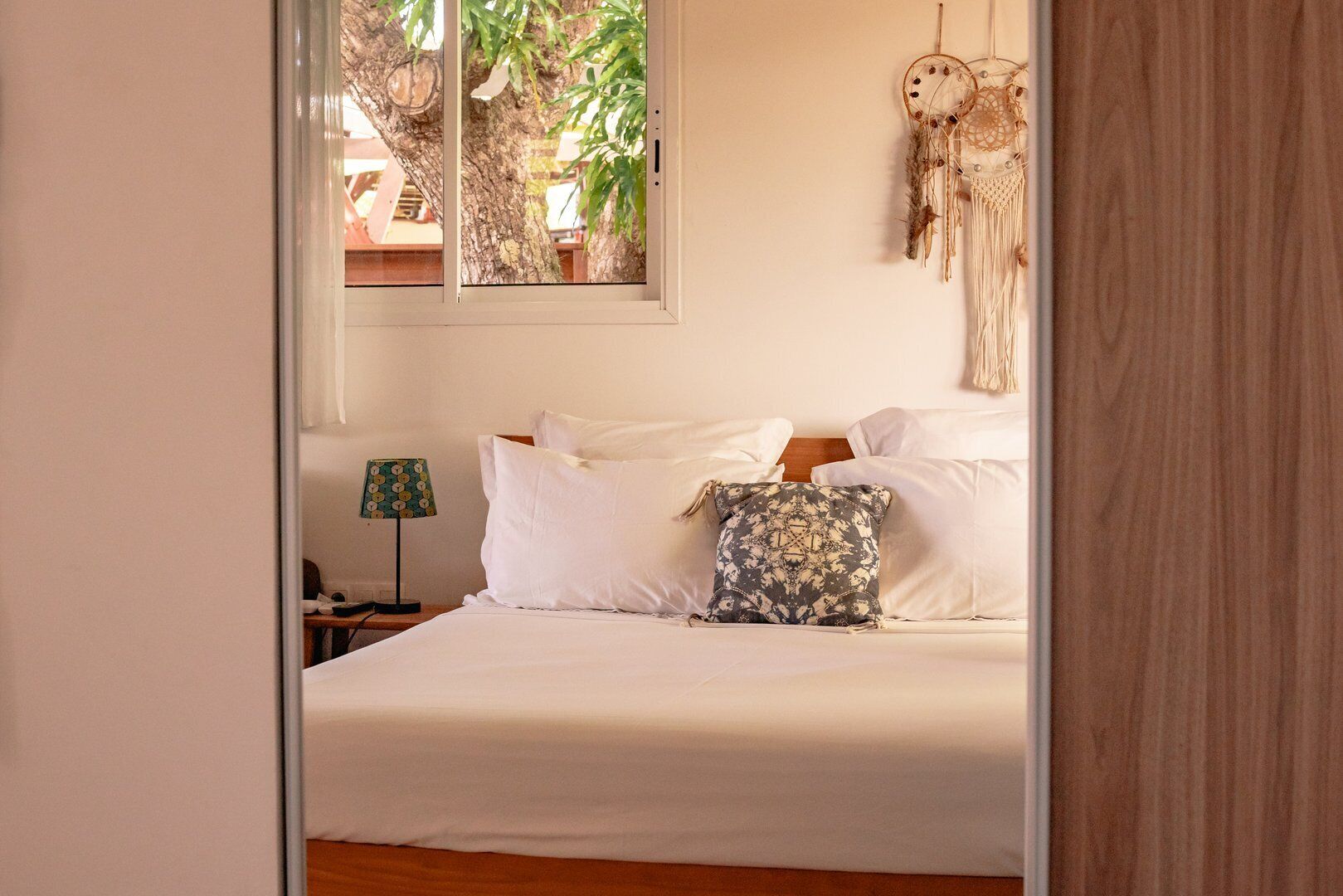 Un dormitorio con cama, almohadas blancas, una almohada estampada, un atrapasueños y una lámpara.