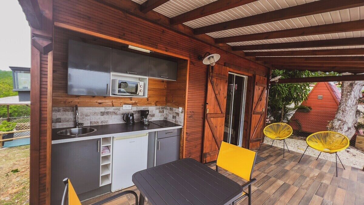 Cocina exterior techada con microondas, pileta y cocina, rodeada de paredes de madera y sillas amarillas.
