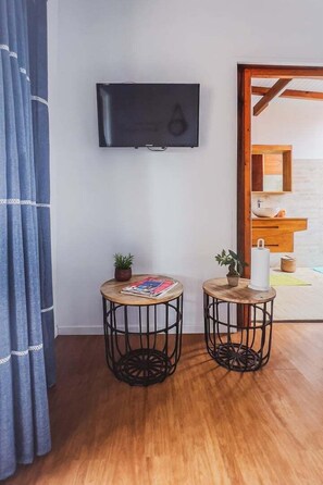 Una habitación con una televisión de pantalla plana montada en la pared, dos mesas redondas con estructura metálica, una pequeña planta en maceta sobre cada mesa y suelo de madera.