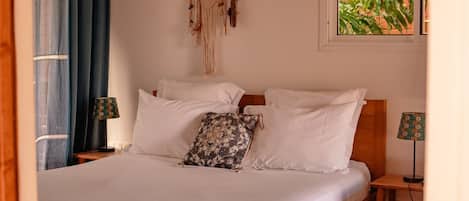 Un lit bien fait, avec une literie blanche, et un attrape-rêves suspendu au-dessus. Deux tables de chevet, chacune avec une lampe, se trouvent de part et d’autre du lit.