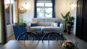 Ein Essbereich mit Tisch, Stühlen und einem Drahtkorb voller Früchte. Im Hintergrund befindet sich eine Couch und ein Fenster.