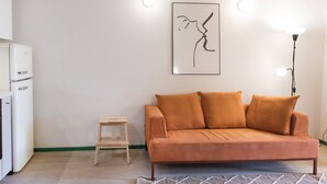 A living room with a sofa, a small table, a refrigerator, and a wall-mounted artwork. - N256 - Numana, brand new three-room apartment with garden (Sirolo)